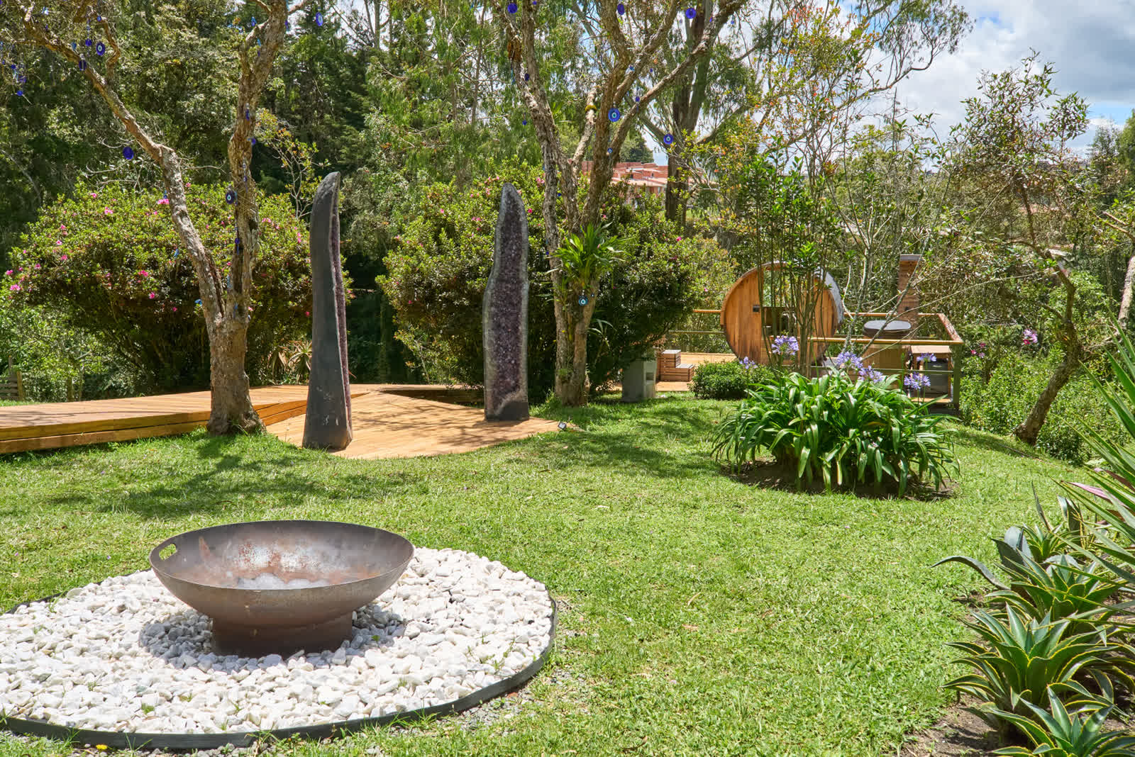 Jardín de EYOWA con geodas de amatista, fogata sobre piedras blancas, sauna de barril y deck de madera entre la vegetación